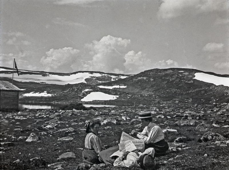 Tre damer på tur i høyfjellet. Sannsynligvis er motivet fra et fjellparti i nærheten av Røldal eller Haukeli. Damene slapper av i lyngen, og en av dem ser også ut til å ha sovnet, med avisen liggende over seg.Fotograf: Thea Larsen, MHB-F.007809, Karmsund folkemuseums fotosamling.