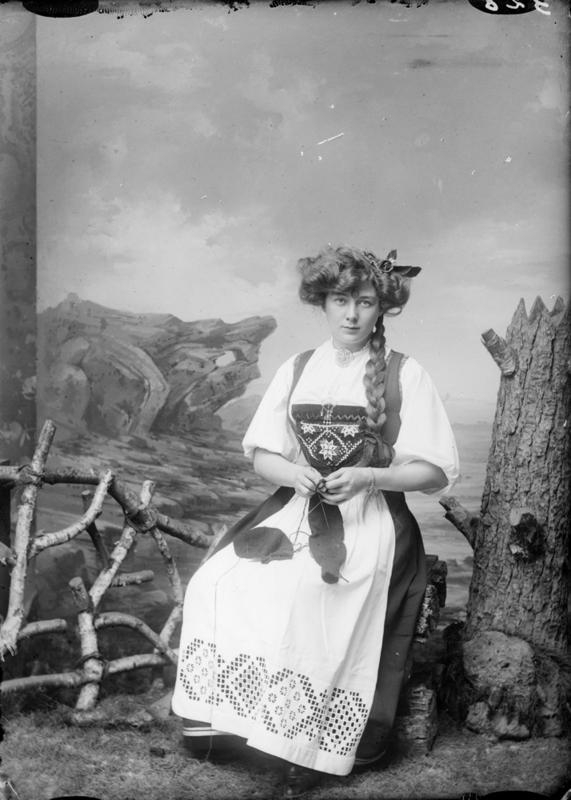 Atelierfotografi av Borghild Hammer. Hun sitter og strikker med bunad på. Rekvisitter som skal gi inntrykk av å være på fjellet. Cirka 1890-1910. Foto: E. H. Torjusen, DFF-EHT0521.